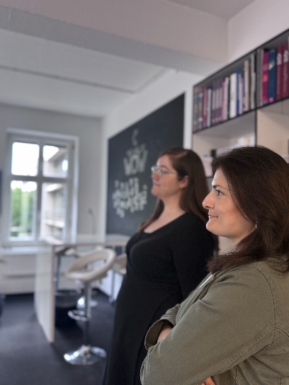 Zwei Frauen in einem Raum mit Büchern und Tafel.
